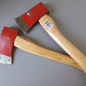 May include: Two axes with wooden handles and red painted heads. The axe heads have a silver blade. The handles are light brown and the axe heads are red. The axes are laying on a grey surface.