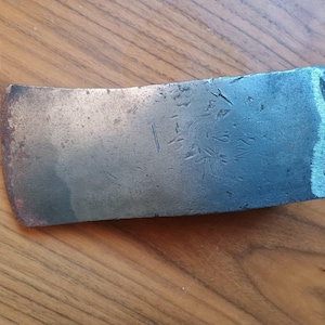 May include: A close-up of a weathered, gray metal axe head with Japanese characters inscribed on the surface. The axe head has a slightly curved shape and a worn edge.