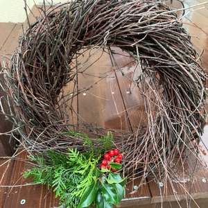 May include: A twig wreath with a natural brown color, decorated with green holly and red berries. The wreath is sitting on a wooden surface.