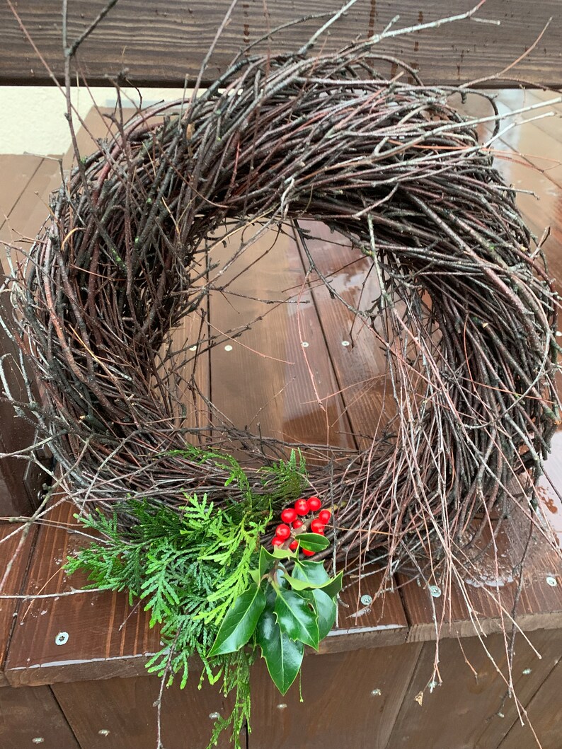 May include: A twig wreath with a natural brown color, decorated with green holly and red berries. The wreath is sitting on a wooden surface.