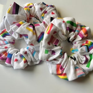 May include: Two white scrunchies with a rainbow pride flag pattern and small red hearts.