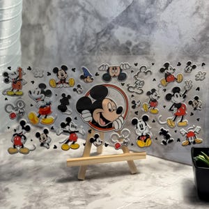 May include: A clear acrylic panel with multiple Mickey Mouse illustrations. The panel is displayed on a small wooden easel. Mickey Mouse is shown in various poses, using red, yellow, black, and white. The background is a grey, marbled effect.