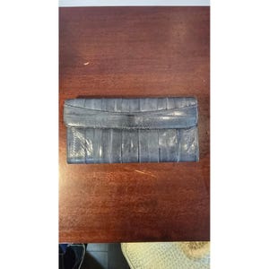 May include: A blue-grey leather clutch wallet with a flap closure. The wallet has a textured, ribbed design and appears to be made of eel skin. The wallet is rectangular and lies flat on a wooden surface.
