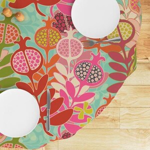 Op de afbeelding: Een ronde tafelkleed met een kleurrijk patroon van gestileerde granaatappels en bladeren. Het tafelkleed is turquoise, roze, rood, oranje en groen.