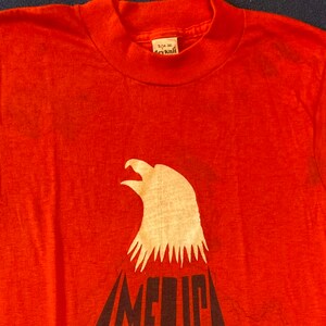 May include: Red t-shirt with a white eagle graphic and the word "AMERICA" printed in black below the eagle. The shirt has a tag that reads "3/24-36" and "Acyknit".