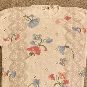 May include: A cream-colored knitted sweater with a floral pattern in shades of pink, blue, and green. The sweater has a crew neck and a relaxed fit.