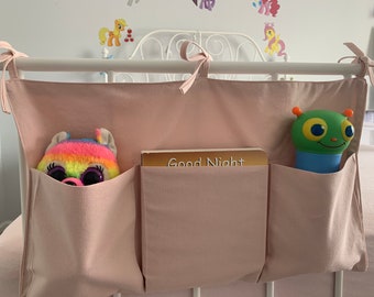 May include: A pink fabric organizer with three pockets hanging on a white metal bed frame. The pockets contain a rainbow stuffed animal, a book titled "Good Night", and a green and blue toy.