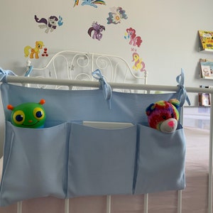 May include: A light blue fabric organizer with three pockets hangs on a white metal bed frame. A green toy with large eyes is in the left pocket, and a rainbow-colored stuffed animal is in the right pocket.