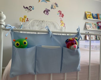 May include: A light blue fabric organizer with three pockets hangs on a white metal bed frame. A green toy with large eyes is in the left pocket, and a rainbow-colored stuffed animal is in the right pocket.