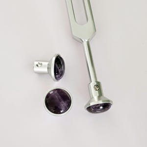May include: A silver tuning fork with two amethyst crystal attachments. The tuning fork has a handle and a tines.
