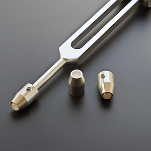 May include: A silver tuning fork with two cylindrical weights. The tuning fork has a U-shaped top and a long handle. The weights are silver with a light-colored, circular end. The items are on a dark surface.