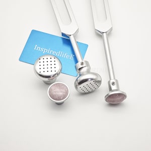 Può includere: Due diapason in metallo color argento con estremità arrotondate e perforate, insieme a due piccoli accessori rotondi separati, uno con superficie perforata e l'altro con un inserto in pietra rosa pallido, e una carta blu con la scritta InspiredlifeE.