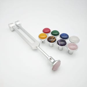 May include: A silver tuning fork with a pink gemstone at the base, accompanied by eight gemstone knobs in various colors. The knobs have silver bases and are arranged in a group. The gemstones include red, green, yellow, brown, blue, white, purple, and pink.
