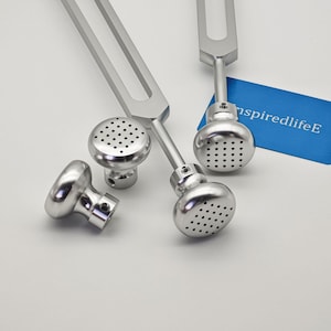Puede incluir: Dos diapasones de metal plateado con extremos pesados que presentan una cuadrícula de pequeñas hendiduras circulares. Los extremos pesados son redondeados y metálicos. Se observa una tarjeta rectangular azul con el texto blanco inspiredlifeE.