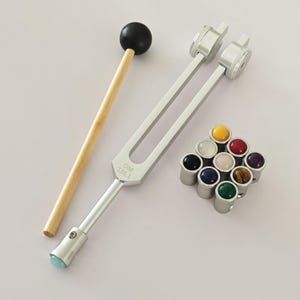 Puede incluir: Un diapasón plateado con un mazo de madera y un conjunto de 12 pequeñas piedras redondas y de colores. El diapasón está etiquetado como "T-136.1 OM".