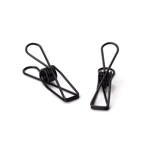 Könnte beinhalten: Zwei schwarze, federbelastete Wäscheklammern aus Metall. Die Wäscheklammern bestehen aus einem einzigen Draht, der zu einer Form gebogen ist, die einem 'S' ähnelt.
