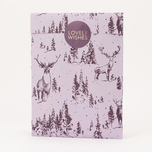 Könnte beinhalten: Eine lavendelfarbene Geschenktüte mit einem sich wiederholenden Muster aus Hirschen und Kiefern. Ein runder Aufkleber mit der Aufschrift "LOVELY WISHES" befindet sich mittig auf der Vorderseite. Goldene Punkte sind über das Design verstreut.