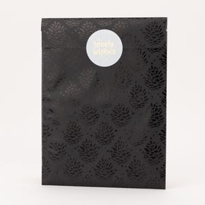 Könnte beinhalten: Eine schwarze Geschenktüte mit einem sich wiederholenden Blumenmuster. Ein weißer runder Aufkleber mit den Worten "lovely. wishes" ist auf der Vorderseite angebracht. Die Tasche ist rechteckig und hat ein glänzendes Finish.