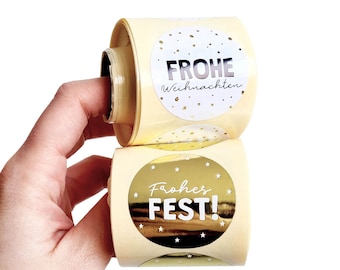 Puede incluir: Dos rollos de pegatinas redondas de color crema. Un rollo tiene las palabras "FROHE Weihnachten" en letras plateadas con confeti dorado. El otro rollo tiene las palabras "Frohes FEST!" en letras blancas con papel dorado y estrellas.