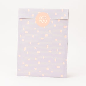 Könnte beinhalten: Eine helllila Geschenktüte mit einem weißen runden Aufkleber, auf dem in weißer Schrift "FOR YOU" steht. Die Tüte hat ein hellrosa und weißes gesprenkeltes Muster.