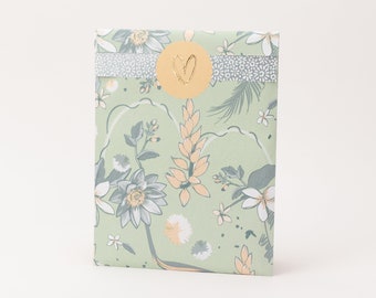 Pode incluir: Um envelope de papel retangular com fundo verde claro e um padrão botânico de flores e folhas cinzentas e cor de pêssego. Uma faixa horizontal com padrão cinzento atravessa o topo, fixada por um autocolante redondo cor de pêssego com um coração dourado.