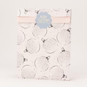 Op de afbeelding: Een cadeauzakje met een lichtroze achtergrond en een patroon van grijze kerstballen. Een lichtblauwe sticker met de tekst "MERRY CHRISTMAS" is aan de zak bevestigd. Een lichtroze lint is ook bevestigd.