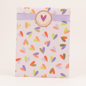 Op de afbeelding: Een rechthoekige geschenkdoos verpakt in lichtpaars papier met een patroon van kleurrijke harten in oranje, groen en paars. Een lichtpaarse band en een ronde sticker met een paars hart en de tekst "wrapped with love".