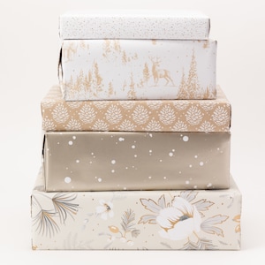 Könnte beinhalten: Ein Stapel von fünf rechteckigen Geschenkboxen, die in verschiedenen dekorativen Papieren verpackt sind. Die Papiere weisen Muster wie goldene Punkte, eine Winterwaldszene mit Hirschen, Tannenzapfenformen, ein Metallic-Finish mit weißen Punkten und florale Designs auf. Die Boxen sind aufeinander gestapelt.
