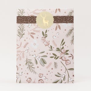 Op de afbeelding: Een rechthoekig cadeaupakket verpakt in lichtroze papier met een botanisch patroon van dennentakken, bessen en kleine sterren. Een donkerbruine horizontale band met witte spikkels loopt over de bovenkant, in het midden met een ronde crèmekleurige sticker.
