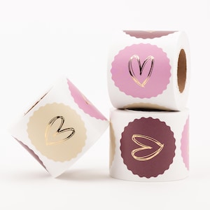May include: Two rolls of adhesive labels featuring scalloped edges and a metallic gold heart design. One roll displays light pink labels, while the other roll features dark maroon labels. The labels are arranged in a stack and a tilted position.