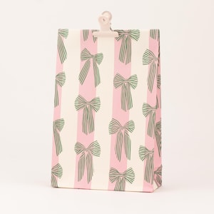 Peut inclure: Un sac cadeau rectangulaire avec un motif répété de nœuds verts sur un fond à rayures roses et blanches. Une pince rose clair est fixée en haut du sac.