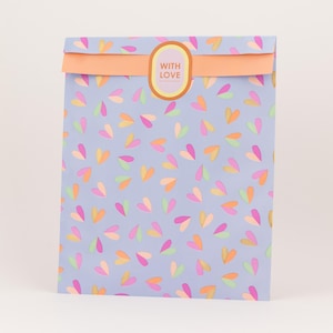Op de afbeelding: Een lichtblauwe cadeauzak met een herhaald patroon van kleurrijke harten in roze, oranje en geel. De tas heeft een gevouwen bovenkant met een oranje band en een ovale sticker met de tekst "WITH LOVE."