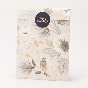 Op de afbeelding: Een cadeauzakje met een bloemenpatroon in crème, grijs en goud. Een donkerblauwe ronde sticker met de tekst "Happy Holidays" staat op de voorkant. De tas heeft een vogel- en bloemontwerp.