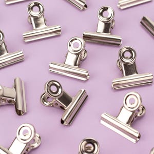 May include: A close-up of silver binder clips on a light purple background. The clips are arranged in a random pattern.