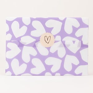 Op de afbeelding: Een cadeau verpakt in lavendelkleurig papier met een patroon van witte harten. Een ronde sticker met een gouden rand en een zwart hart sluit het pakket af. Het papier is netjes gevouwen.