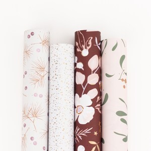 Könnte beinhalten: Vier Rollen dekoratives Geschenkpapier mit verschiedenen floralen und abstrakten Mustern. Designs umfassen goldene Tannennadeln, goldene Sprenkel, weiße Blumen auf bordeauxrotem Hintergrund und grüne Blätter auf rosa Hintergrund.