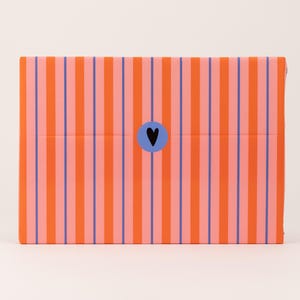 Könnte beinhalten: Eine rechteckige Geschenkbox, verpackt in Papier mit vertikalen Streifen in Orange, Rosa und Blau. Ein blauer Aufkleber mit einem schwarzen Herzen ist mittig auf der Vorderseite angebracht. Die Box ist geschlossen und bereit zum Verschenken.