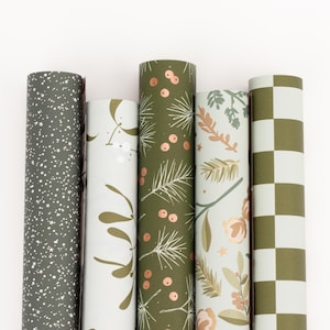 Pode incluir: Cinco rolos de papel de embrulho decorativo. Os designs incluem um padrão salpicado cinza escuro, folhas brancas e douradas, agulhas de pinheiro e bagas verdes, padrões florais e um tabuleiro de xadrez verde e branco.
