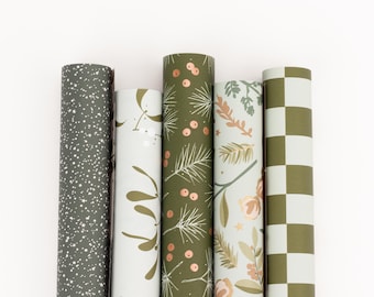 Könnte beinhalten: Fünf Rollen dekoratives Geschenkpapier. Designs umfassen ein dunkelgraues, gesprenkeltes Muster, weiße und goldene Blätter, grüne Tannennadeln und Beeren, florale Muster und ein grün-weißes Schachbrettmuster.