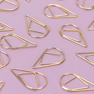Peut inclure: Une collection de trombones dorés en forme de goutte d'eau avec un triangle à l'intérieur. Les trombones sont éparpillés sur une surface rose pâle.