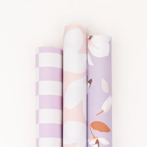 Könnte beinhalten: Drei Rollen dekoratives Geschenkpapier. Die erste Rolle hat ein Schachbrettmuster in Weiß und Lavendel. Die zweite Rolle hat einen rosa Hintergrund mit weißen Blumendesigns. Die dritte Rolle hat einen lavendelfarbenen Hintergrund mit weißen und orangefarbenen Blumendesigns.