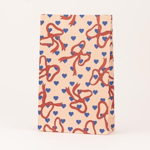 Peut inclure: Un sac cadeau rectangulaire avec un motif répété de nœuds rouges et de cœurs bleus sur un fond pêche clair. Le sac est debout, présentant le motif sur le devant et le côté.