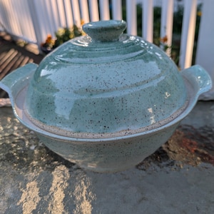 May include: A light green ceramic lidded bowl with a speckled finish and two side handles. The bowl features a rounded base and a matching domed lid with a small knob handle on top. The surface has a glossy glaze with visible dark brown speckles.
