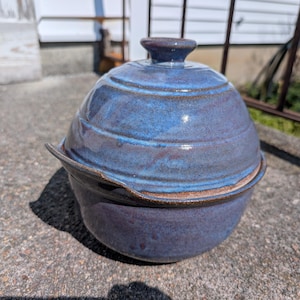 May include: A handmade, blue ceramic casserole dish with a lid. The dish has a rounded shape with a dark blue glaze and a brown rim. The lid has a small handle on top.