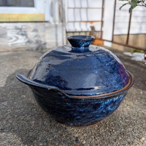 May include: A dark blue ceramic casserole dish with a lid. The dish has a rounded shape with a handle on each side and a small knob on the lid. The dish has a glossy finish and a speckled texture.