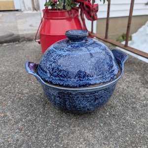May include: A blue ceramic casserole dish with a lid. The dish has a speckled pattern and two handles. The lid has a small knob on top. The dish is sitting on a concrete surface.