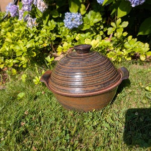 May include: A round, lidded ceramic casserole dish featuring a dark brown and black glaze with horizontal ridged textures. The pot has two small, integrated side handles and a central knob on the lid, showcasing a rustic, hand-thrown appearance.