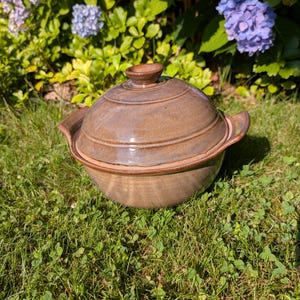 May include: A round, brown ceramic casserole dish with a matching domed lid and two side handles. The pottery features a smooth, glossy glaze with subtle variations in tone, showcasing a functional design with a central knob on the lid.