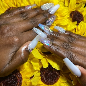 May include: Close-up of long, white acrylic nails with iridescent glitter accents and rhinestones. The nails are displayed against a background of yellow artificial sunflowers.