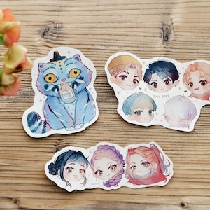 Könnte beinhalten: Eine Sammlung von Aufklebern mit Aquarellillustrationen. Einer zeigt eine blaue Katze mit Hut, während andere stilisierte Porträts von Personen mit verschiedenen Frisuren und Farben zeigen. Die Aufkleber befinden sich auf einer Holzoberfläche.